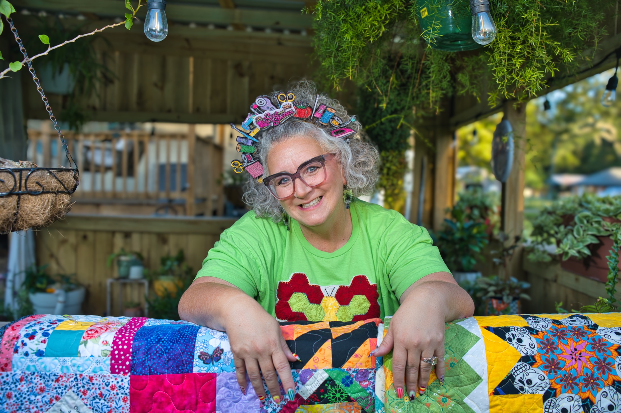 Angela in a flower headscarf, smiling and leaning on a patchwork quilt at her garden pergola